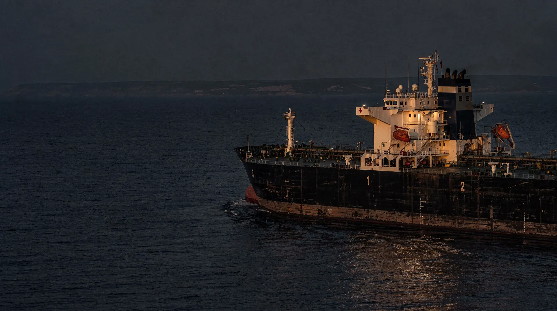 An unmarked oil tanker crosses dark water at dusk beneath a hazy shoreline.