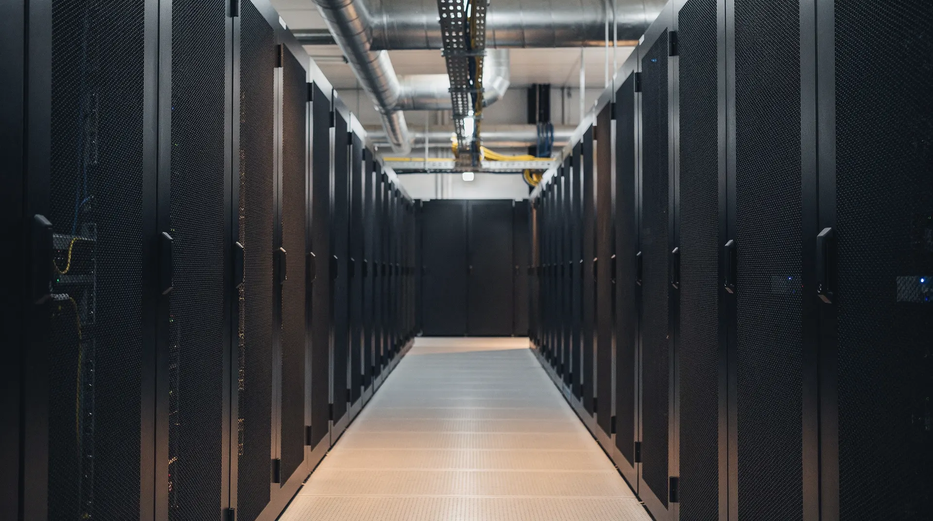 Closed server racks line a data-center aisle beneath overhead cooling pipes.