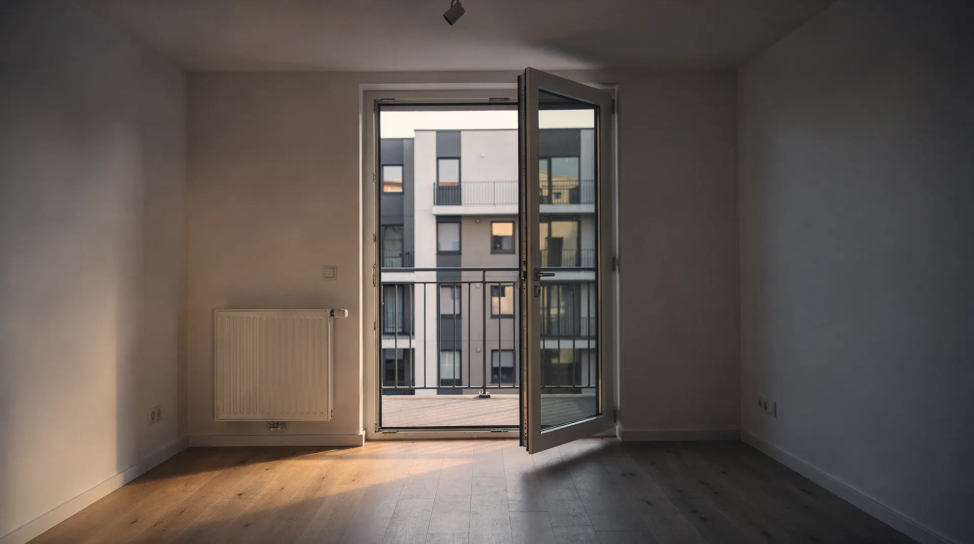 An empty apartment opens onto a balcony overlooking neighboring multifamily buildings.