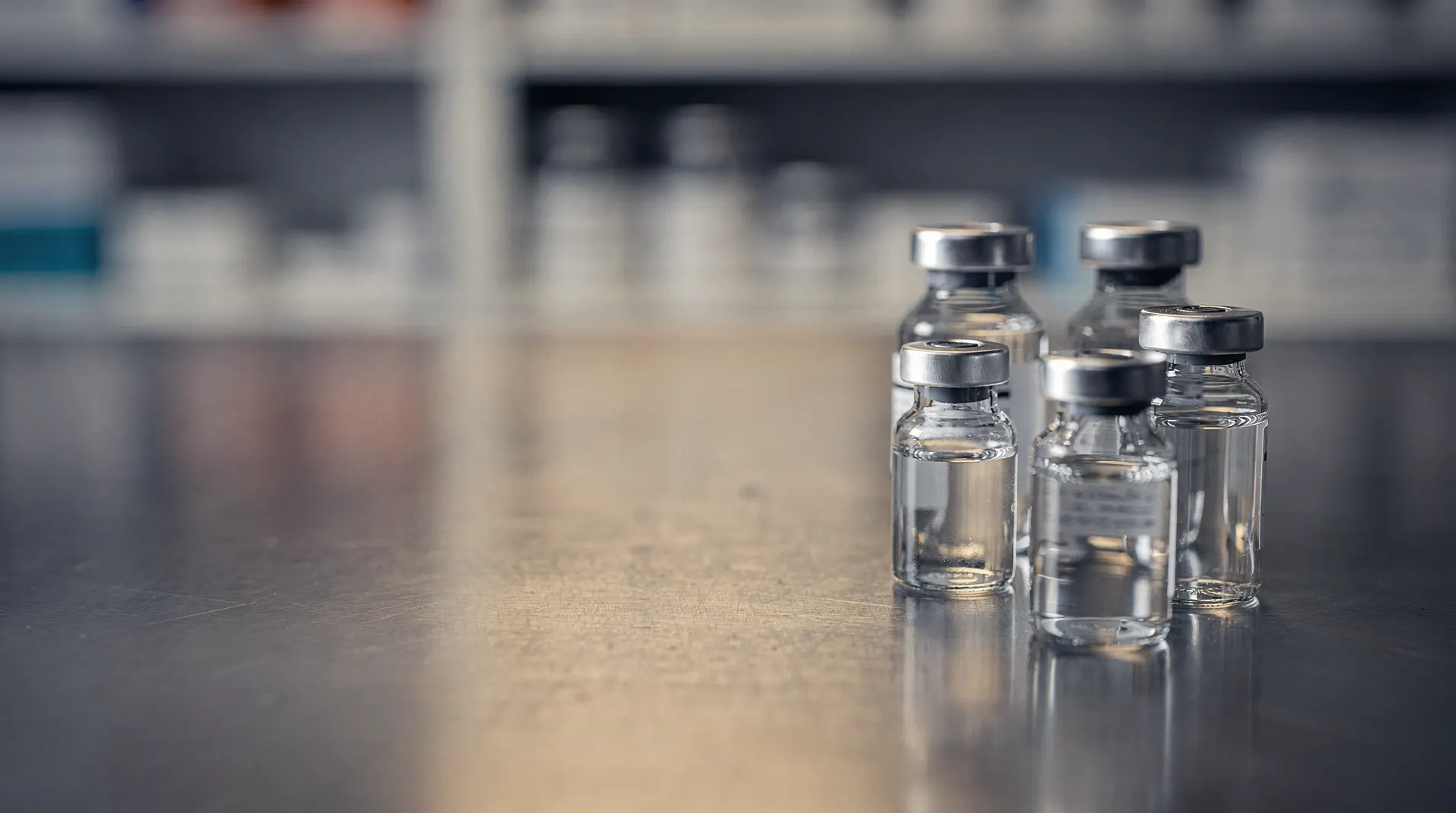 Unlabeled pharmaceutical vials sit on a hospital pharmacy workbench against softly blurred shelves.