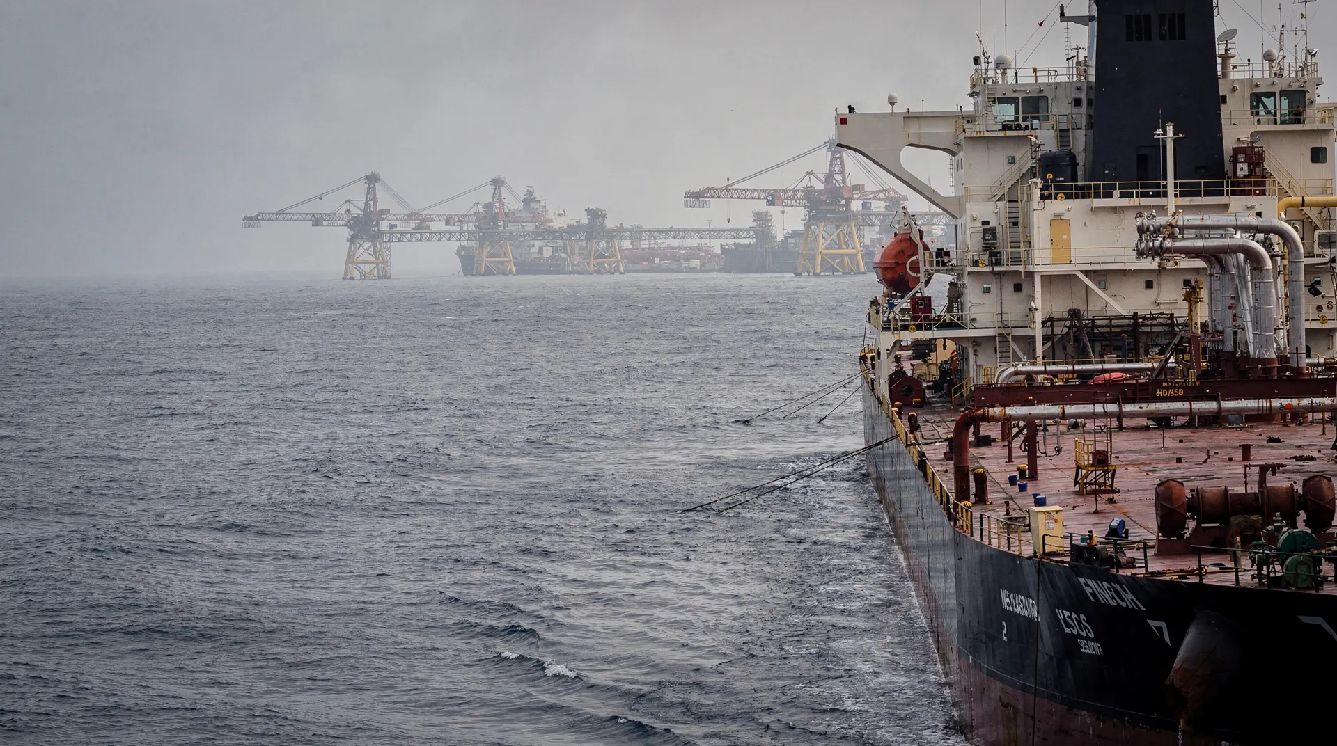 A crude oil tanker passes distant loading gantries in a hazy harbor.
