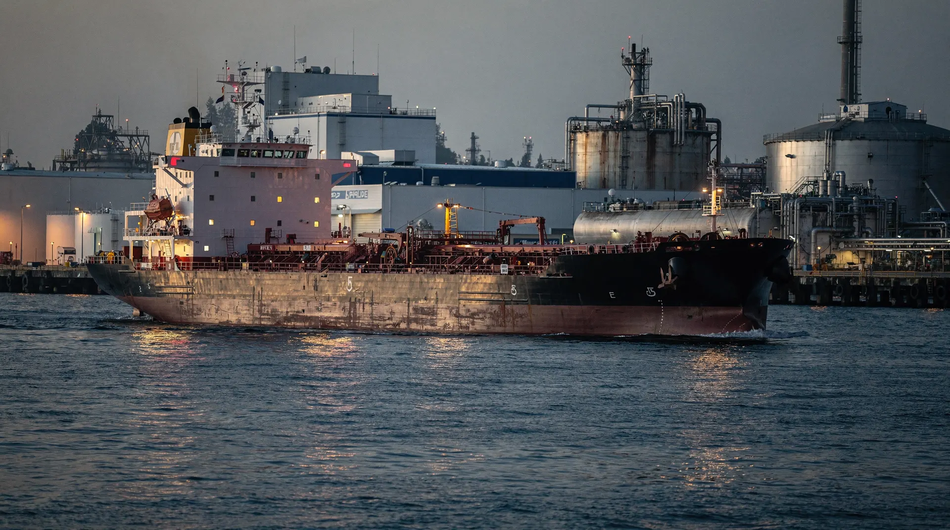 An unmarked diesel tanker approaches a refinery terminal across a harbor at dusk.