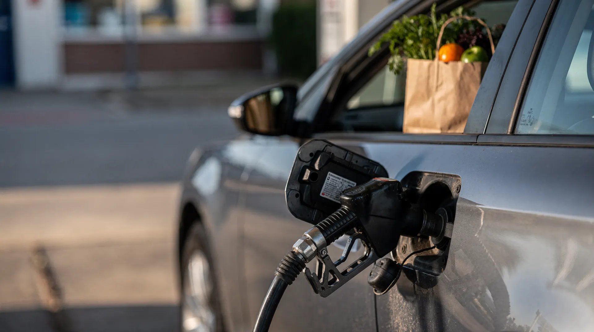 Gasoline nozzle in a car's fuel filler, with a grocery bag softly visible through the window.