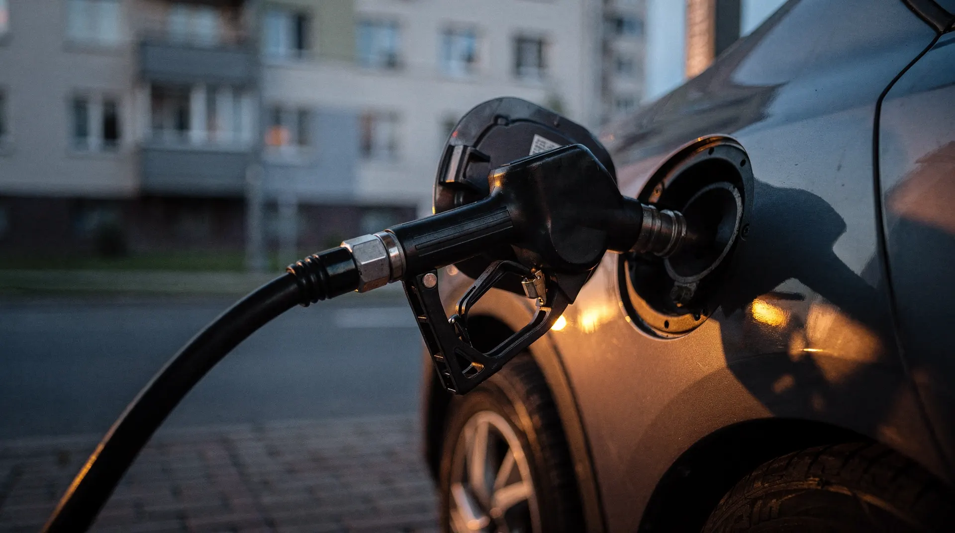 A fuel nozzle fills a car, with an apartment building blurred across the street at dusk.