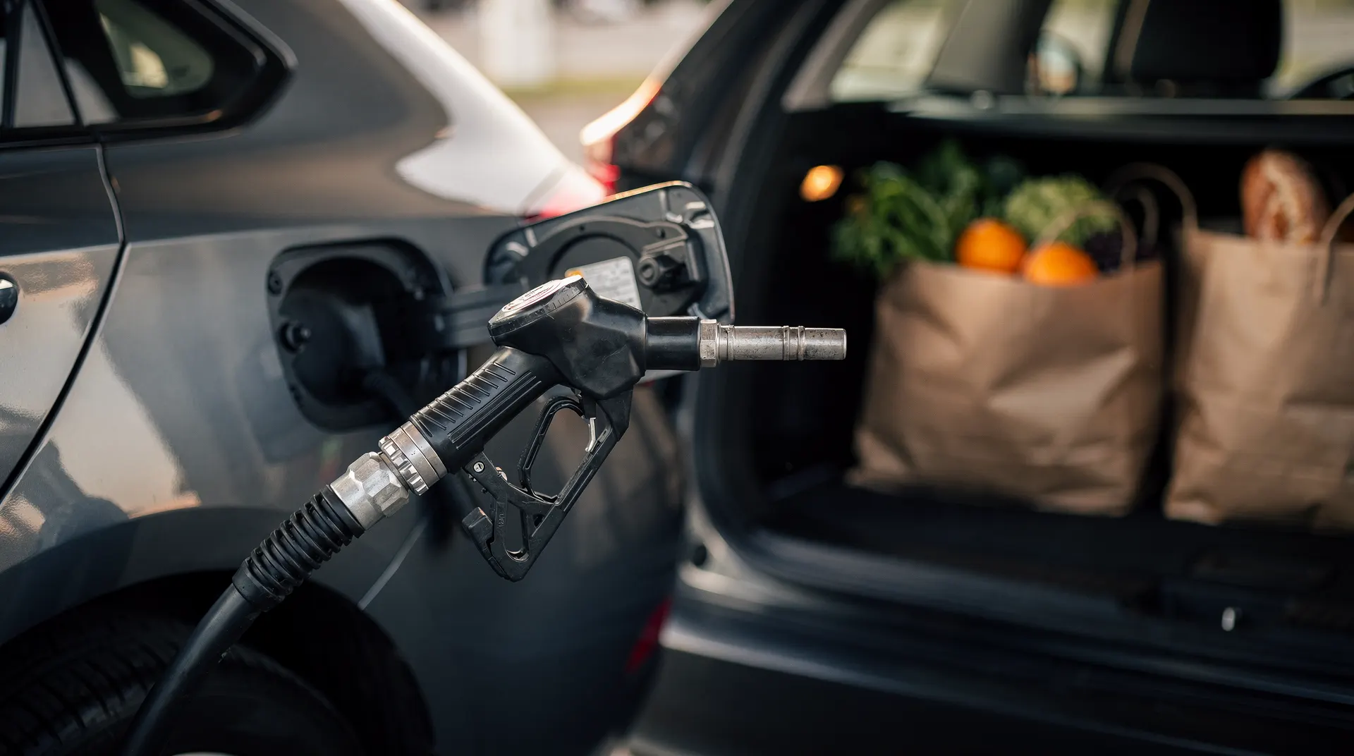 A fuel-pump nozzle fills a car, with bags of groceries visible in its open cargo hatch.