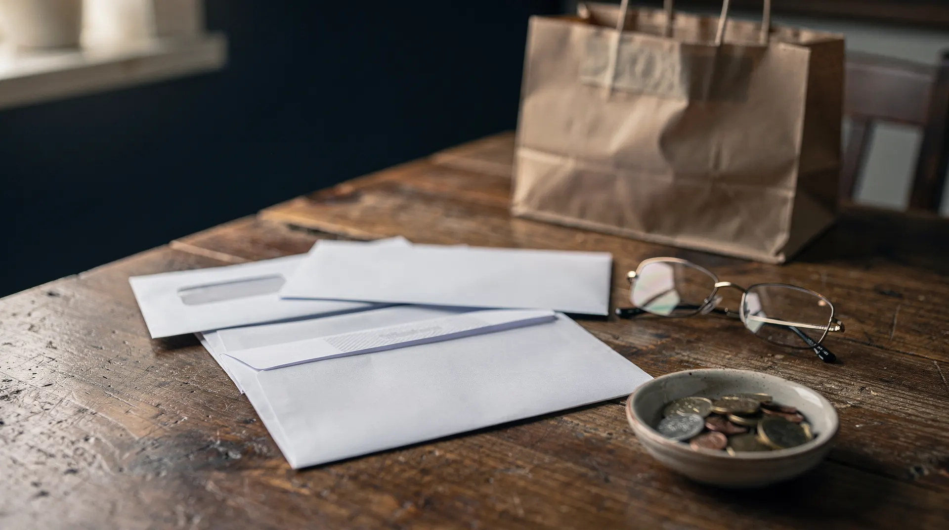 Blank unopened envelopes, reading glasses and a dish of coins on a kitchen table beside a blurred grocery bag.