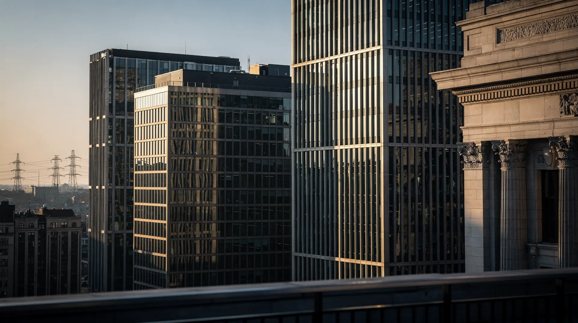 Office towers rise above a quiet street, with a stone-fronted building and distant power pylons.
