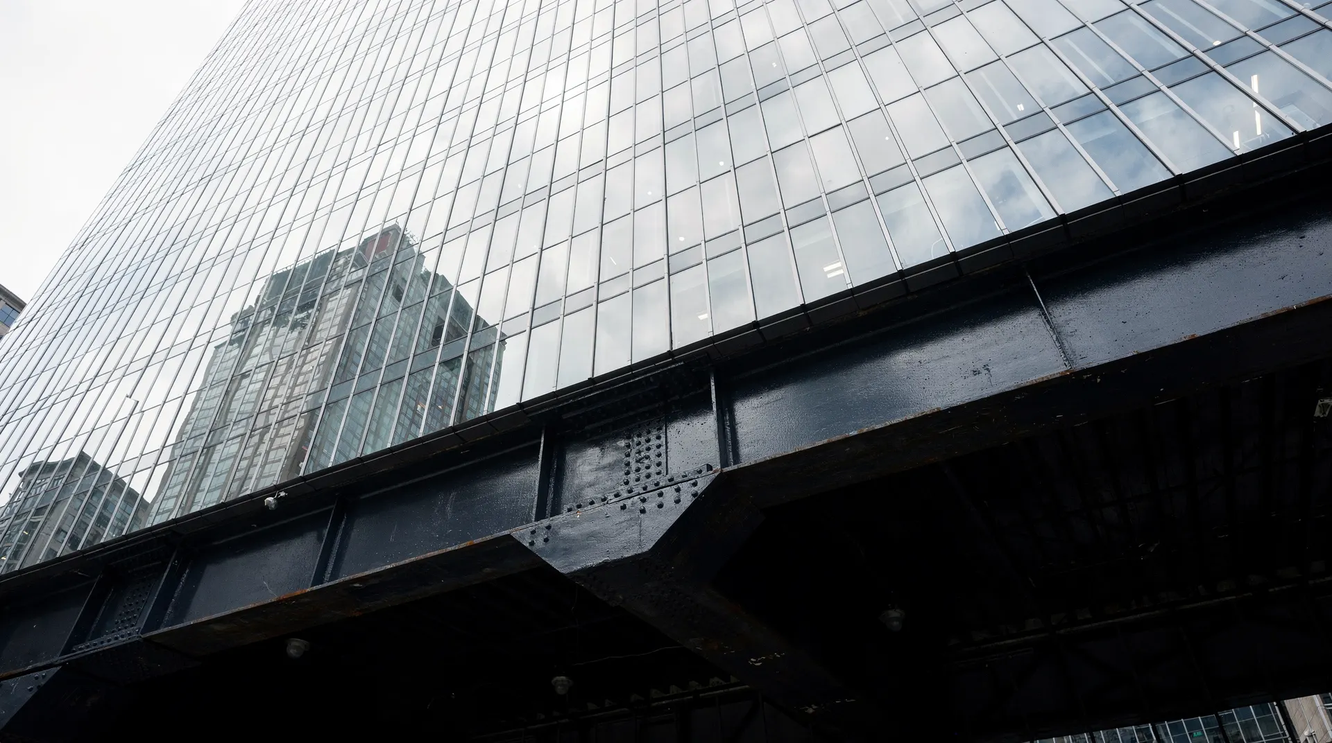 A lone steel girder rises beneath a vast glass skyscraper facade, viewed from the street.