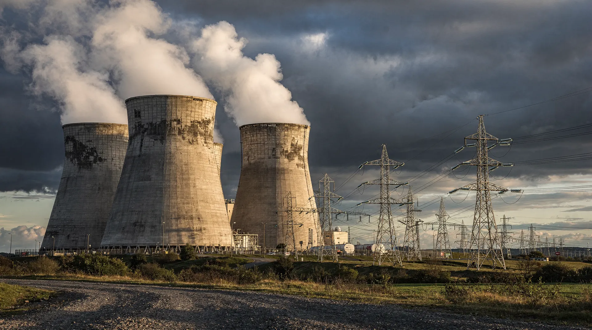Nuclear power station cooling towers stand beside transmission pylons beneath a gathering cloud bank.