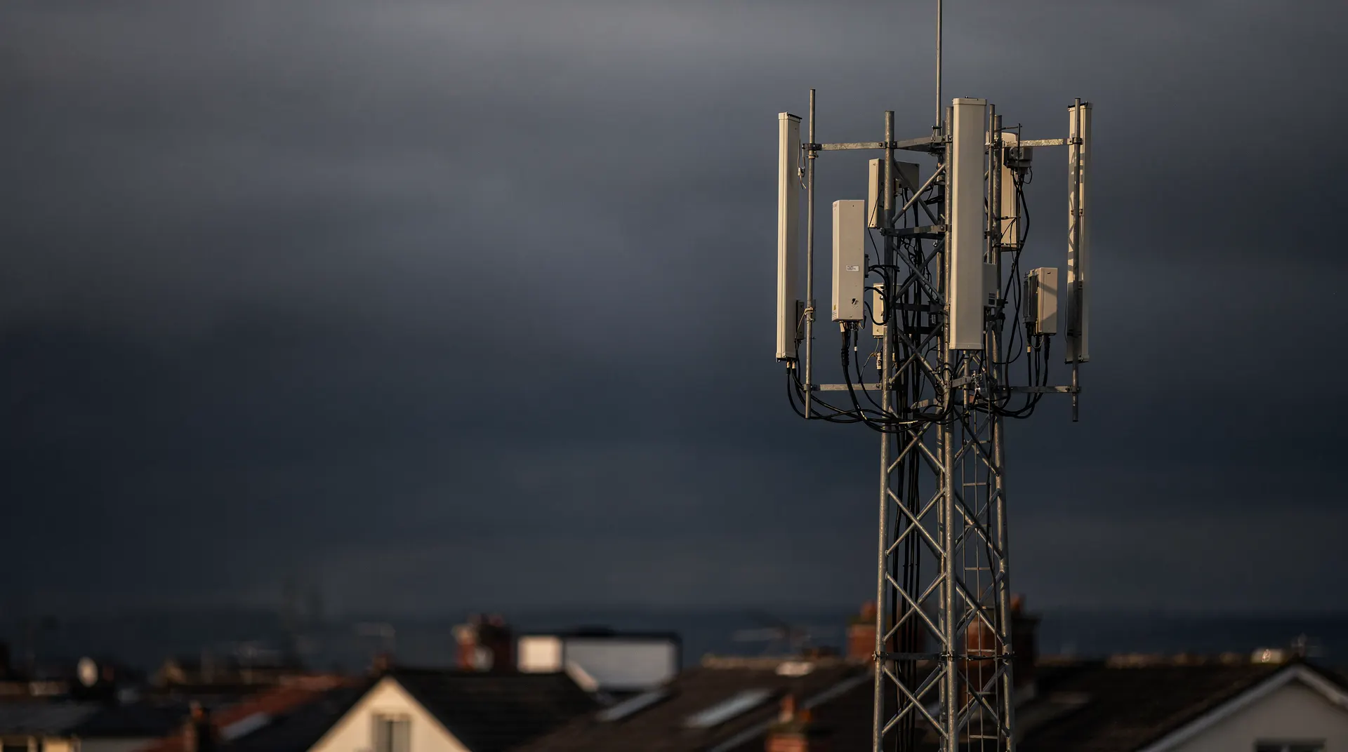 A cellular tower with several antenna arrays rises above distant rooftops under an overcast sky.