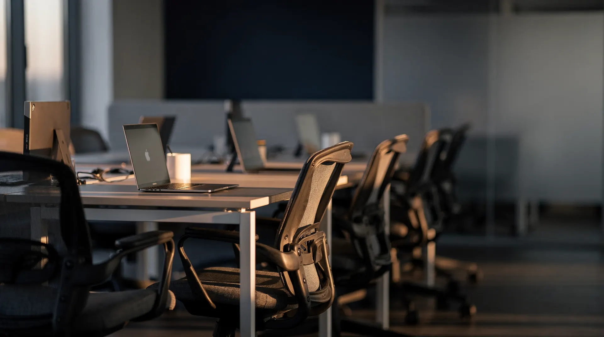 Empty office desks and chairs recede into a quiet workplace in soft dawn light.