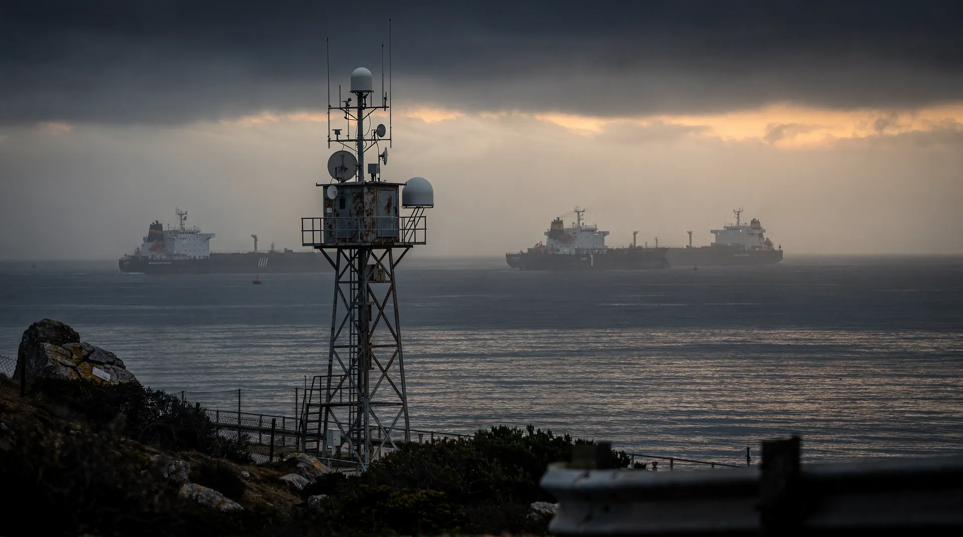 Two unmarked oil tankers sit far apart in a hazy narrow sea passage beneath a heavy sky.