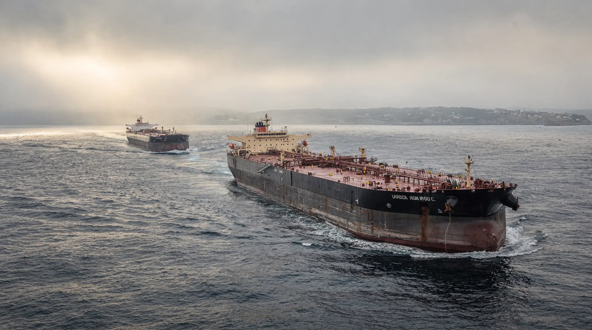 A crude oil tanker crosses a narrow, hazy sea passage with another tanker in the distance.