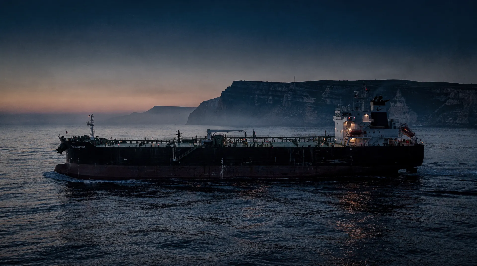 An intact oil tanker sails through open water at dusk, with a rugged coastline in the distance.