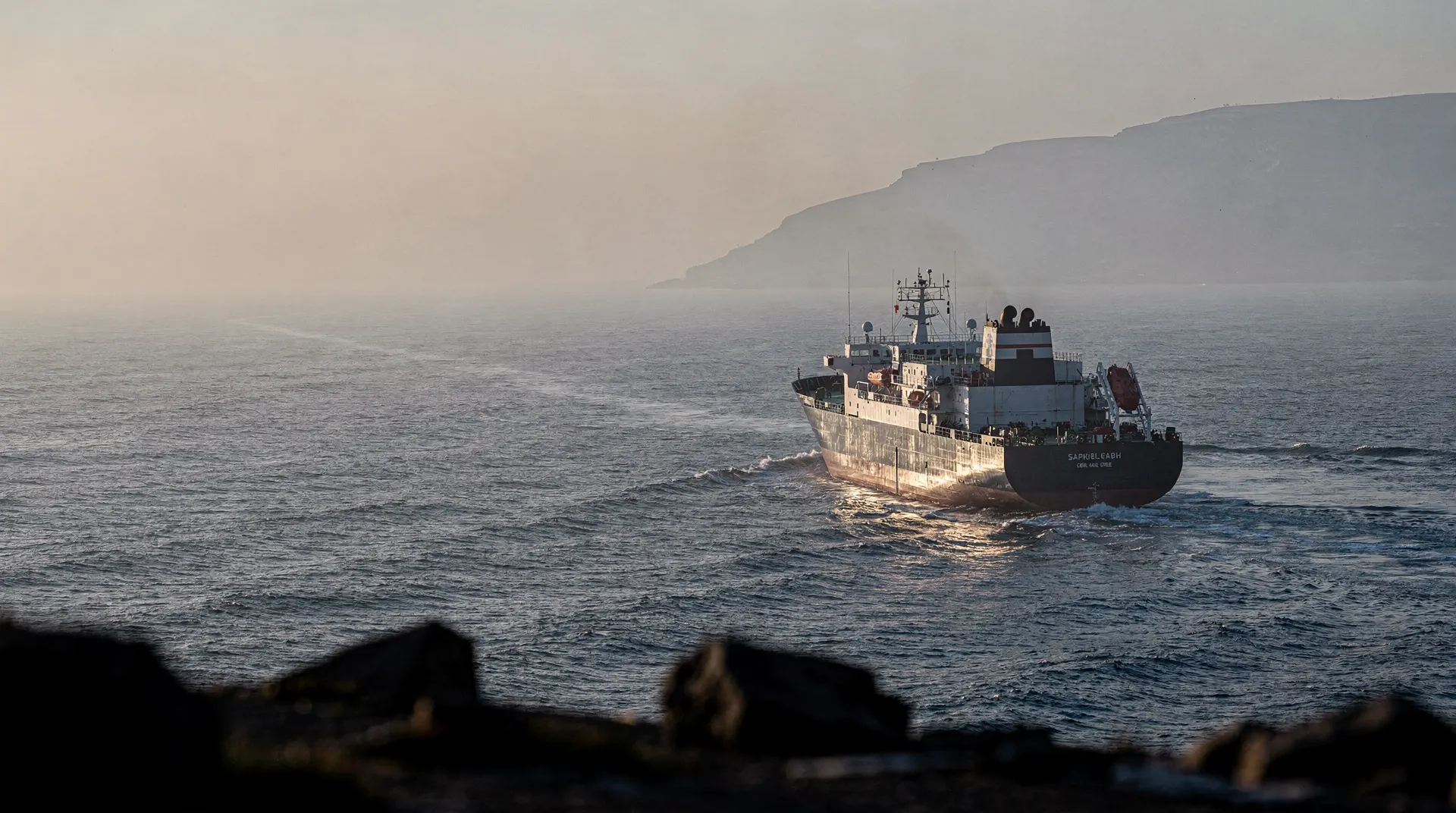 An unmarked merchant vessel crosses hazy water with a distant coastline behind it.