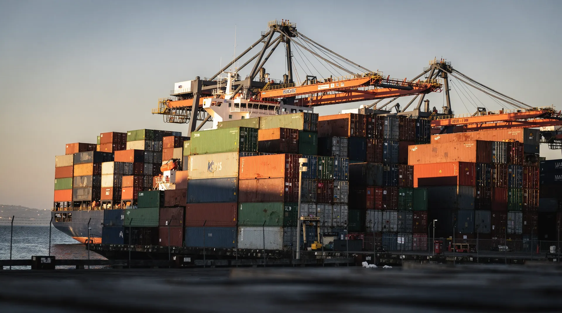 Dense stacks of shipping containers fill a port terminal beside a ship and towering cranes.