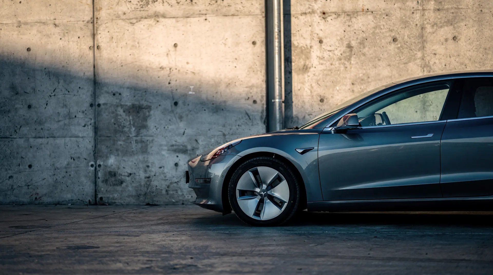 An electric sedan stands beside a dim factory loading bay in late-afternoon light.