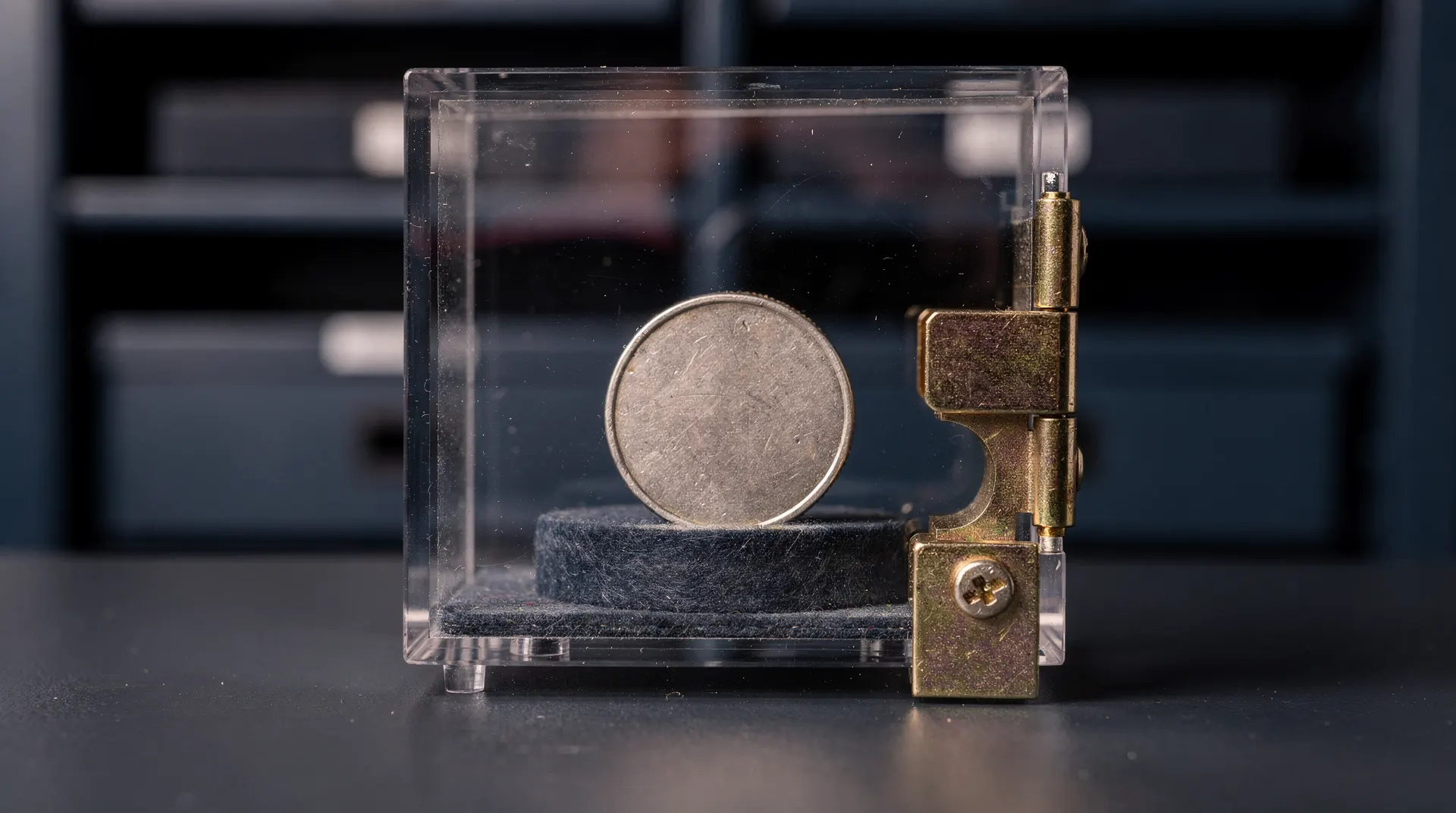 An unmarked metal coin sits inside a locked transparent box against a dark vault backdrop.