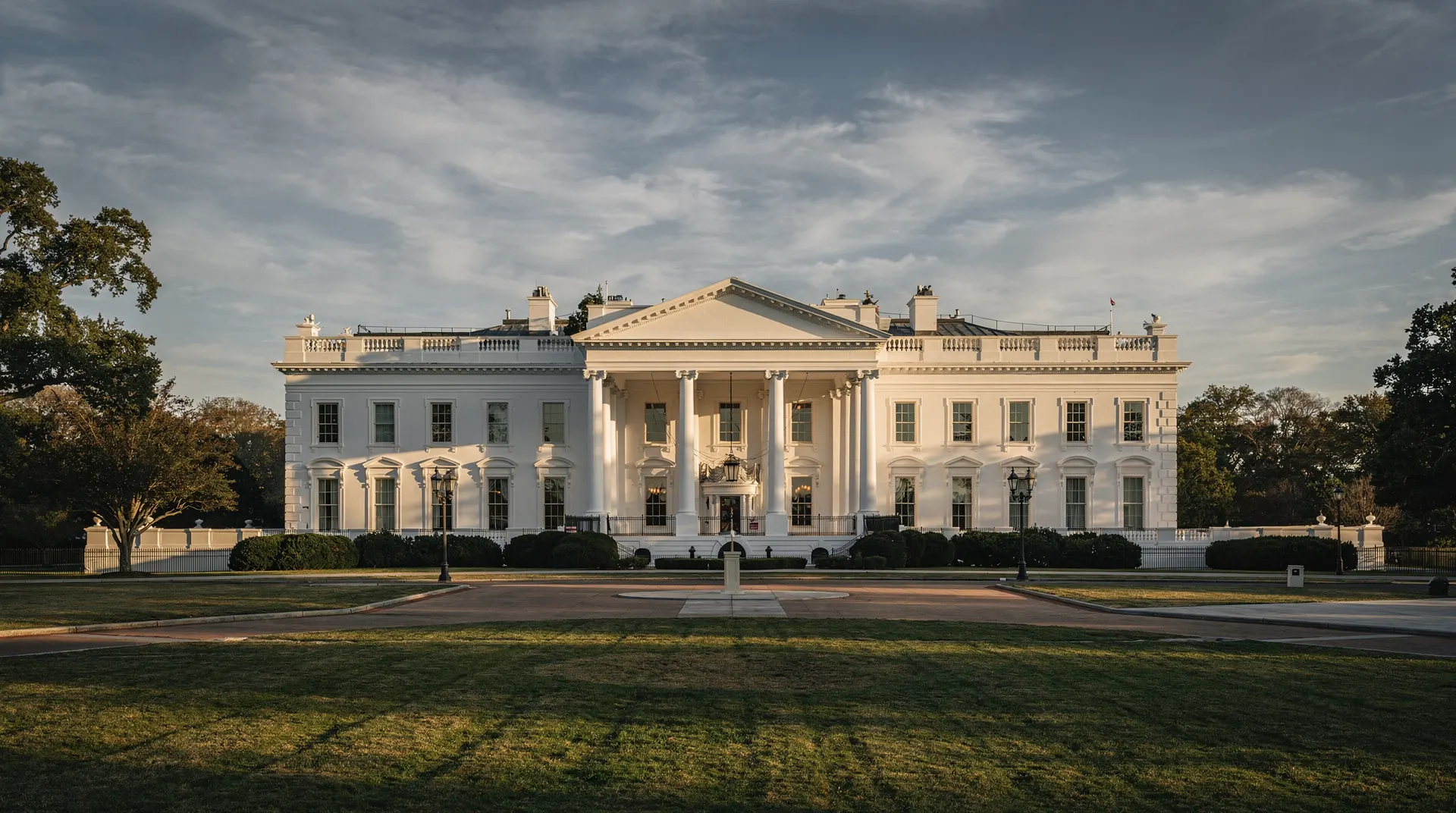 The White House stands beyond empty grounds under a blue-gray sky in late-afternoon light.