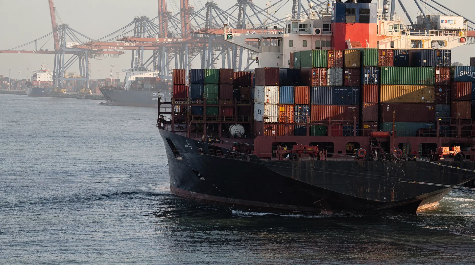 A loaded container ship approaches a crowded port terminal, with cranes and a waiting vessel beyond.