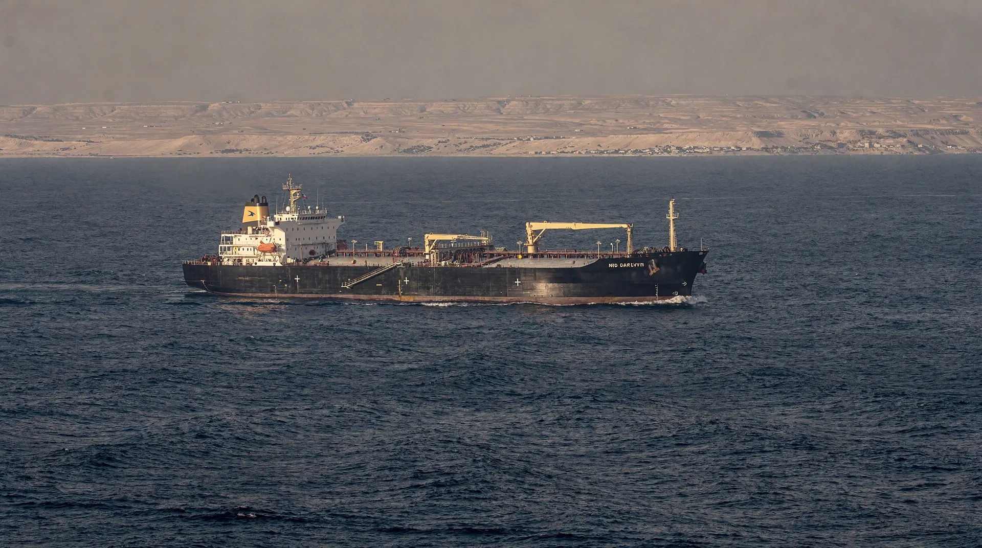 An unmarked oil tanker crosses open water with an arid coastline on the hazy horizon.