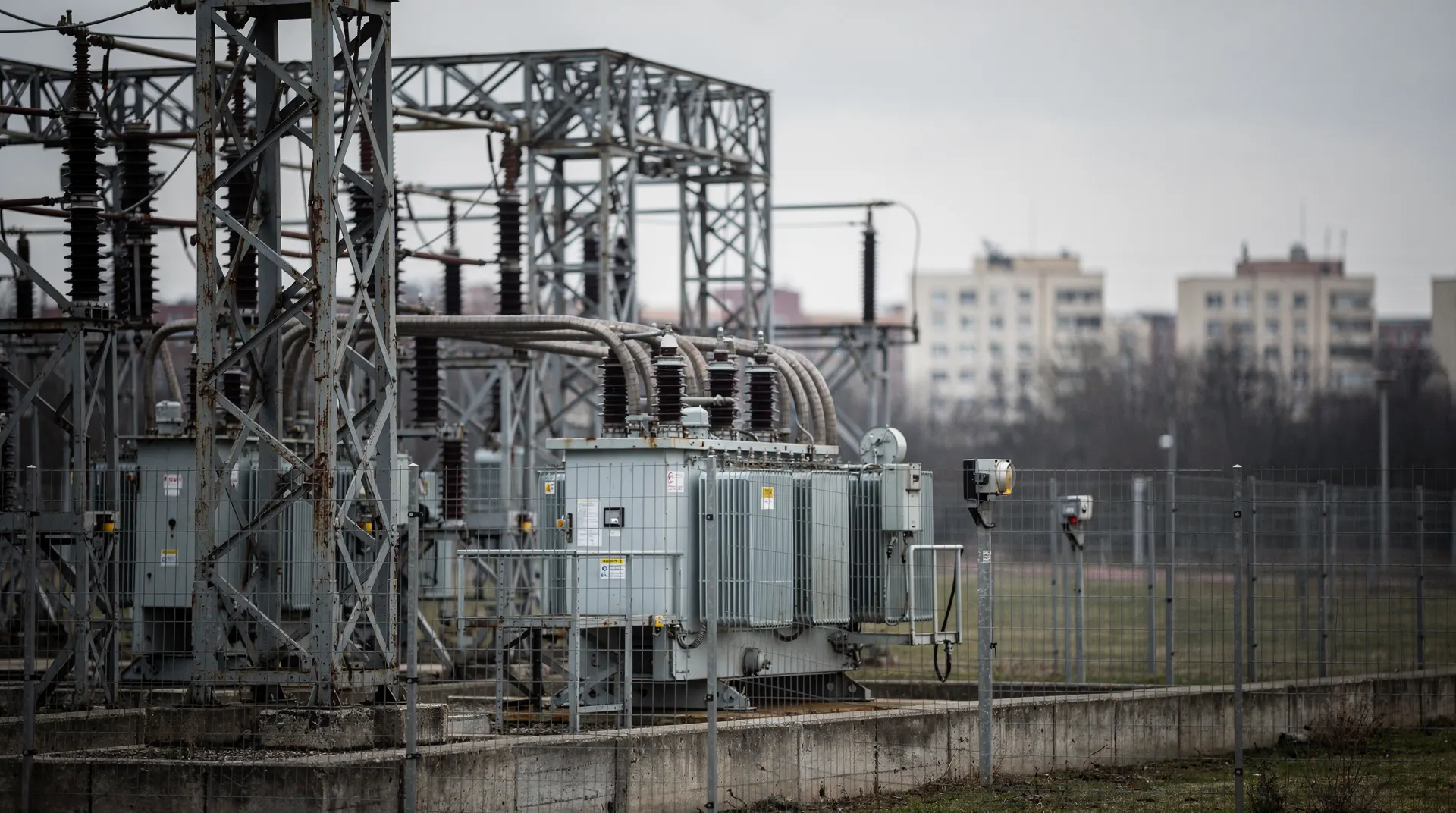 Electrical substation equipment stands in front of distant apartment buildings beneath an overcast sky.