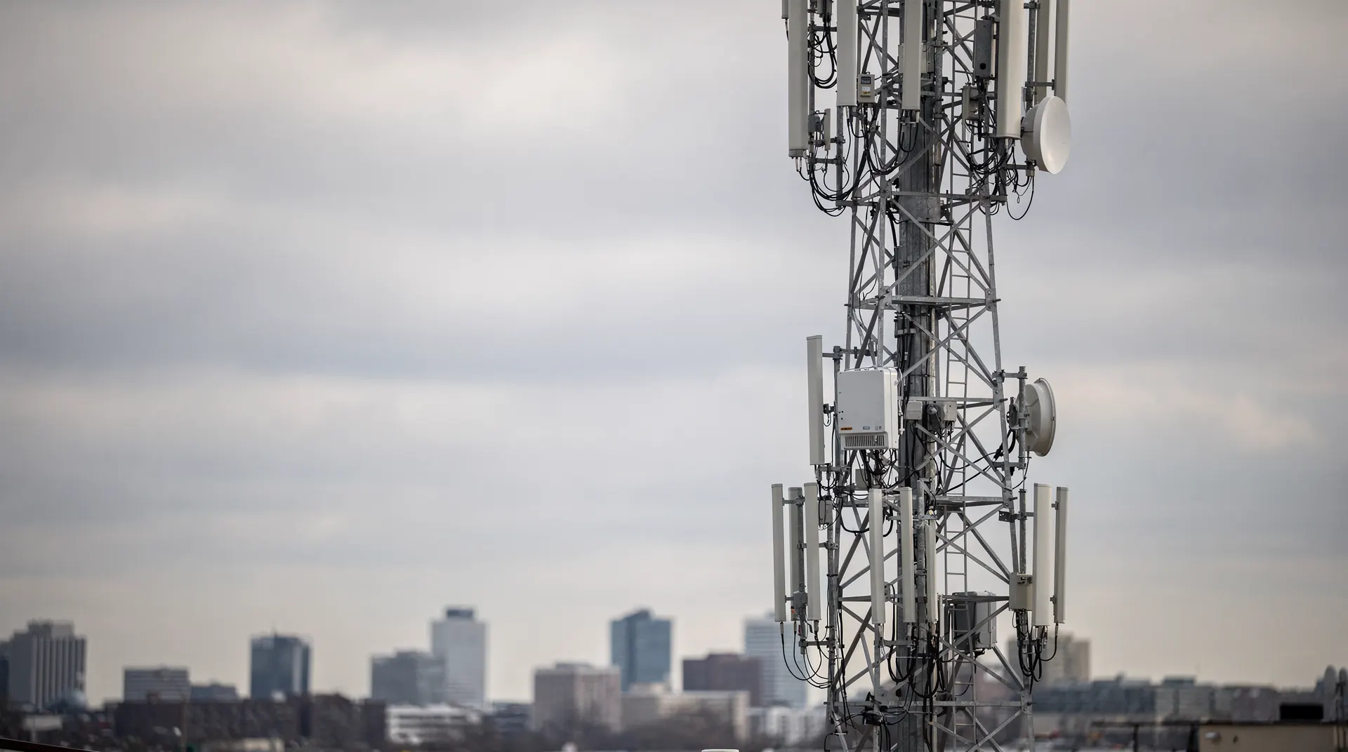 Cellular antennas on a tower beneath heavy clouds, with a blurred city skyline in the distance.