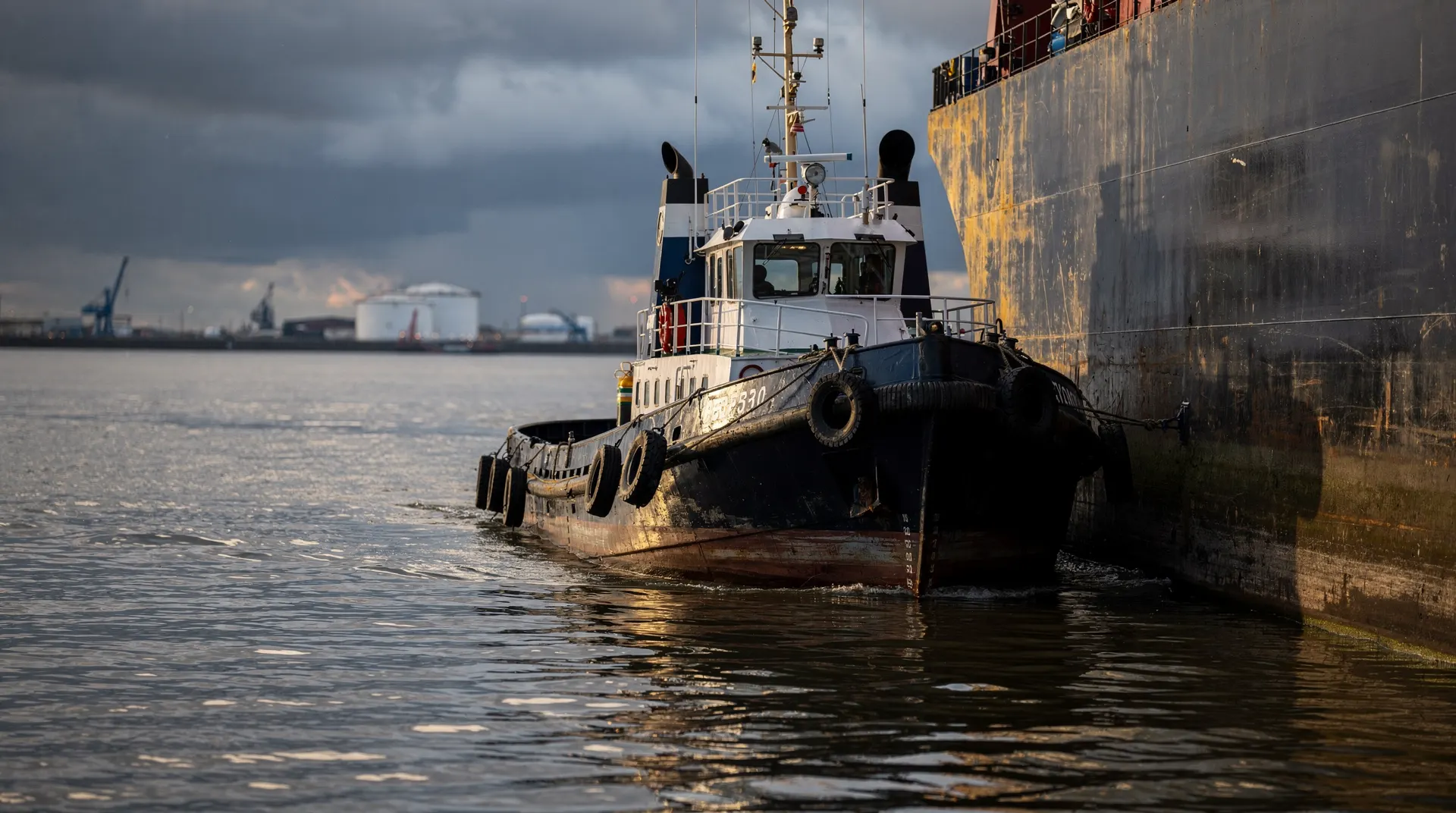 An unmarked oil tanker crosses a shipping channel beneath low storm clouds, with an industrial shoreline in the distance.
