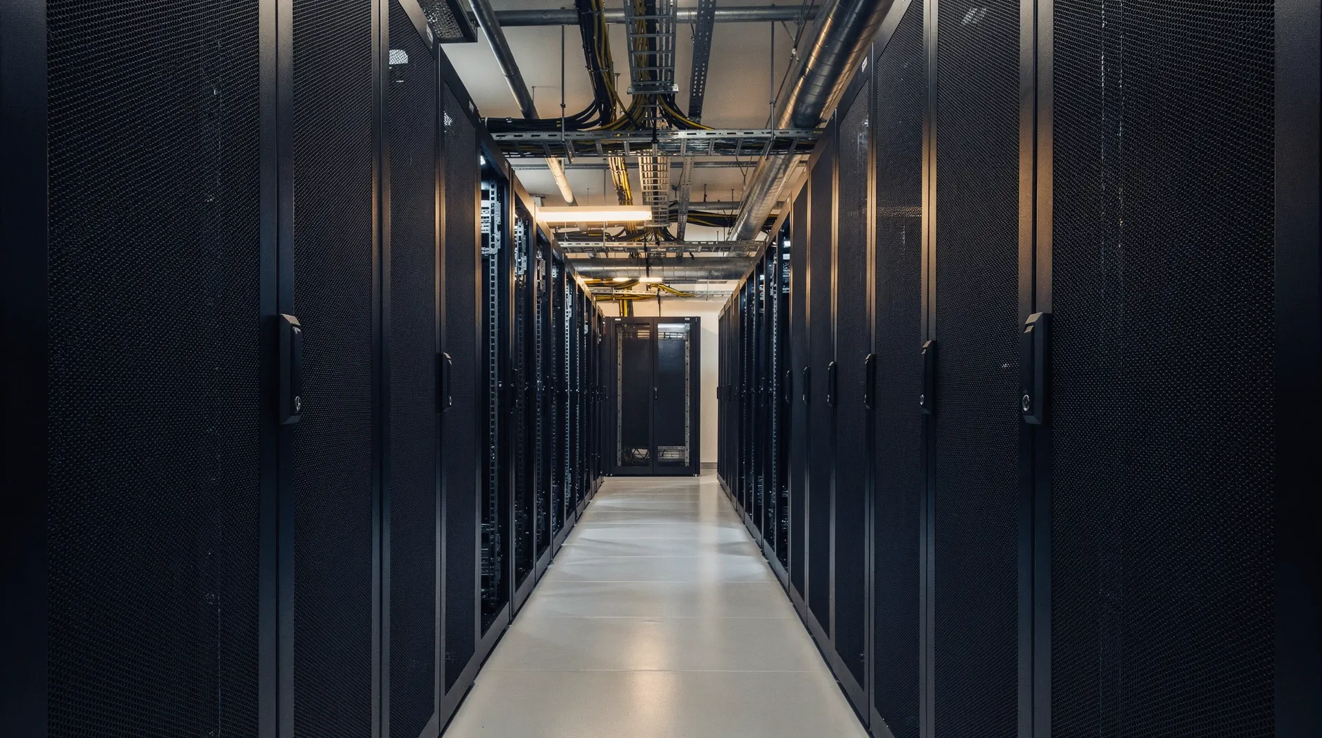 Installed server racks face empty bays in a dim datacenter aisle beneath cooling pipes.