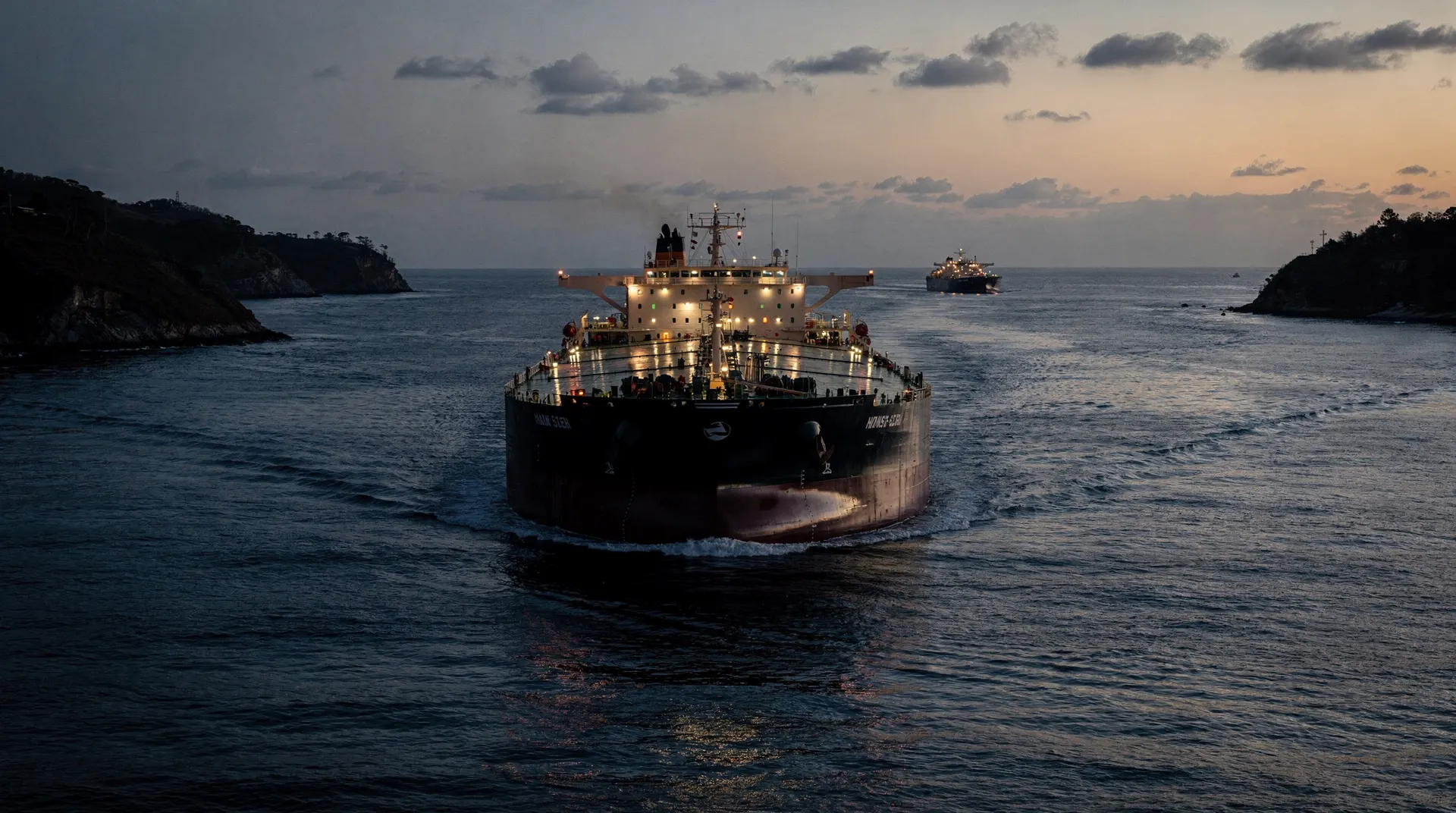 An oil tanker crosses a narrow sea passage at dusk, with another tanker visible in the distance.